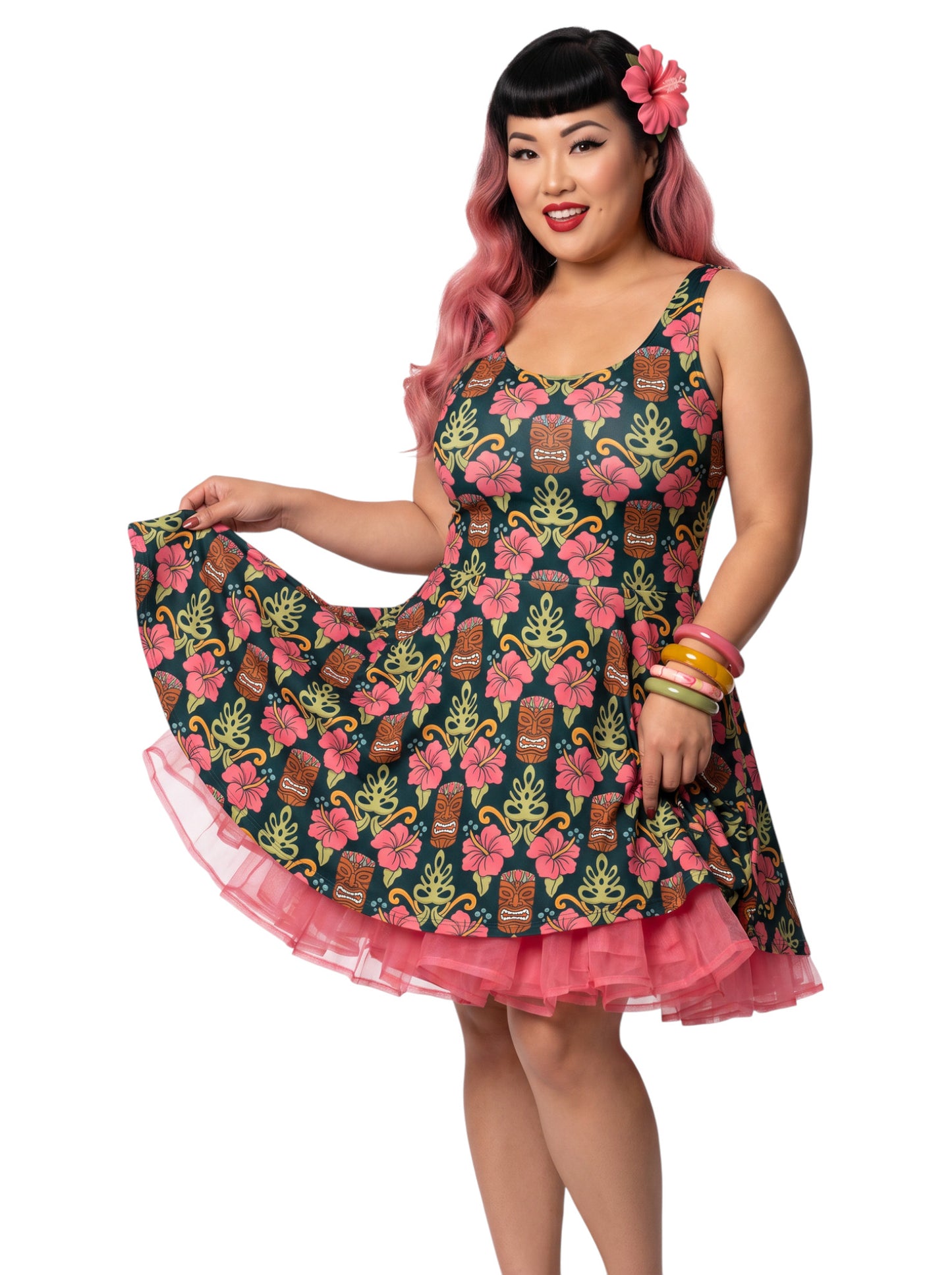 A plus size woman wearing a dark green 1950s-style pinup rockabilly swing dress with an all-over tropical print of tiki masks, pink hibiscus flowers, and monstera leaves. 