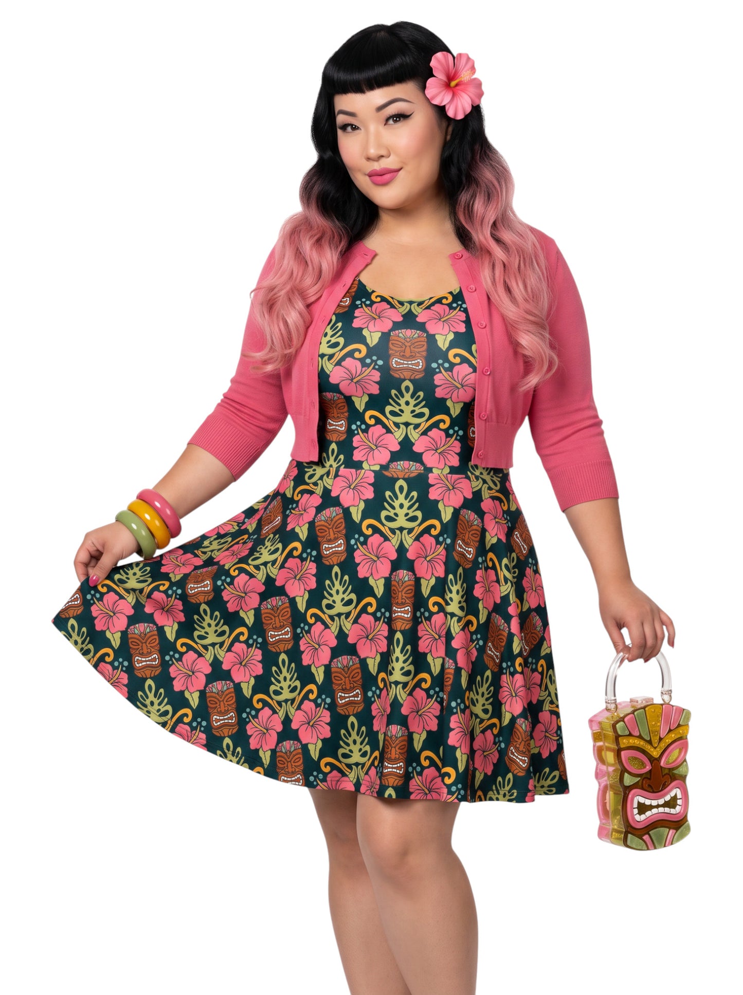 A plus size woman wearing a dark green 1950s-style pinup rockabilly swing dress with an all-over tropical print of tiki masks, pink hibiscus flowers, and monstera leaves. 