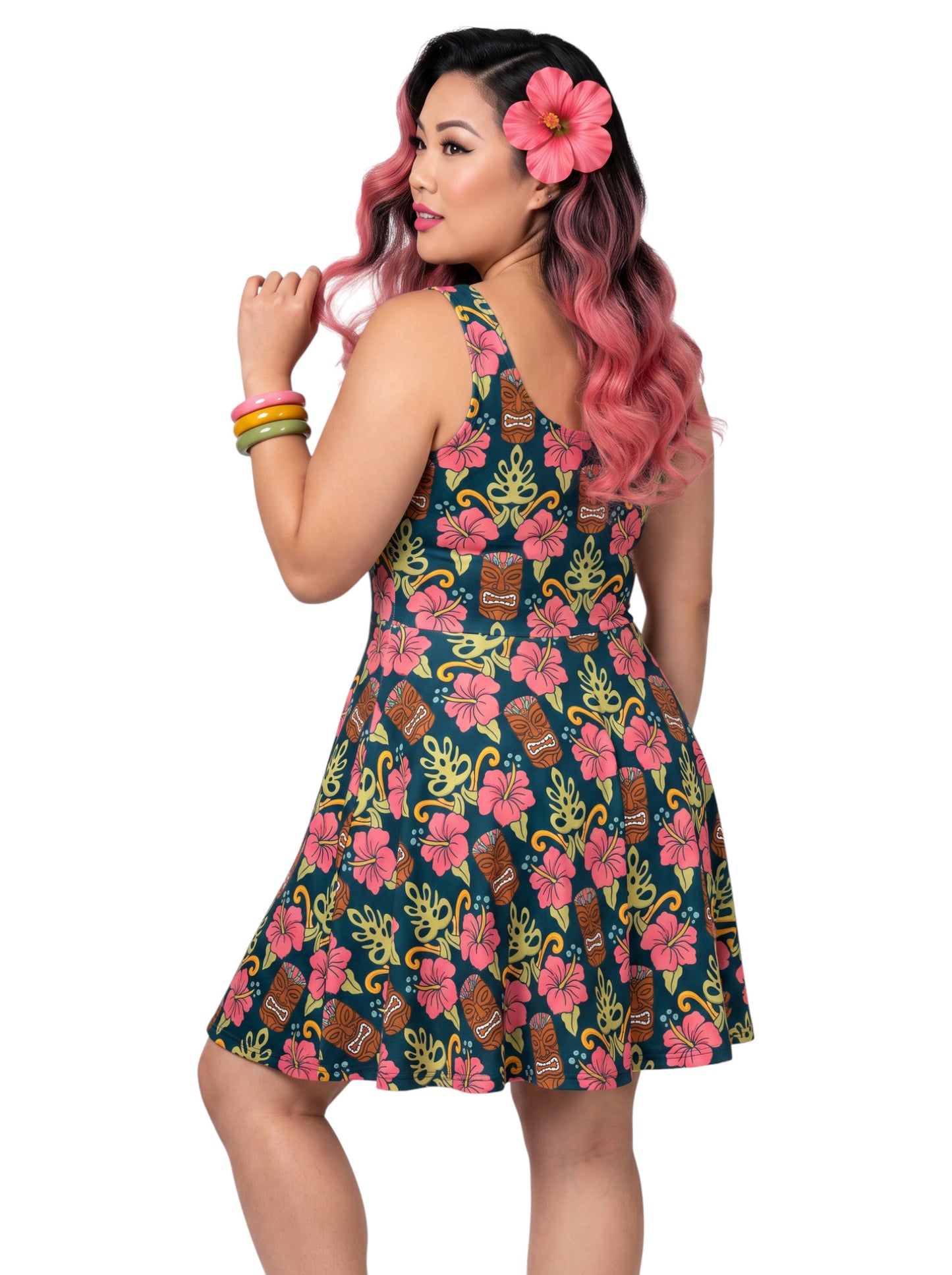 A plus size woman wearing a dark green 1950s-style pinup rockabilly swing dress with an all-over tropical print of tiki masks, pink hibiscus flowers, and monstera leaves. 